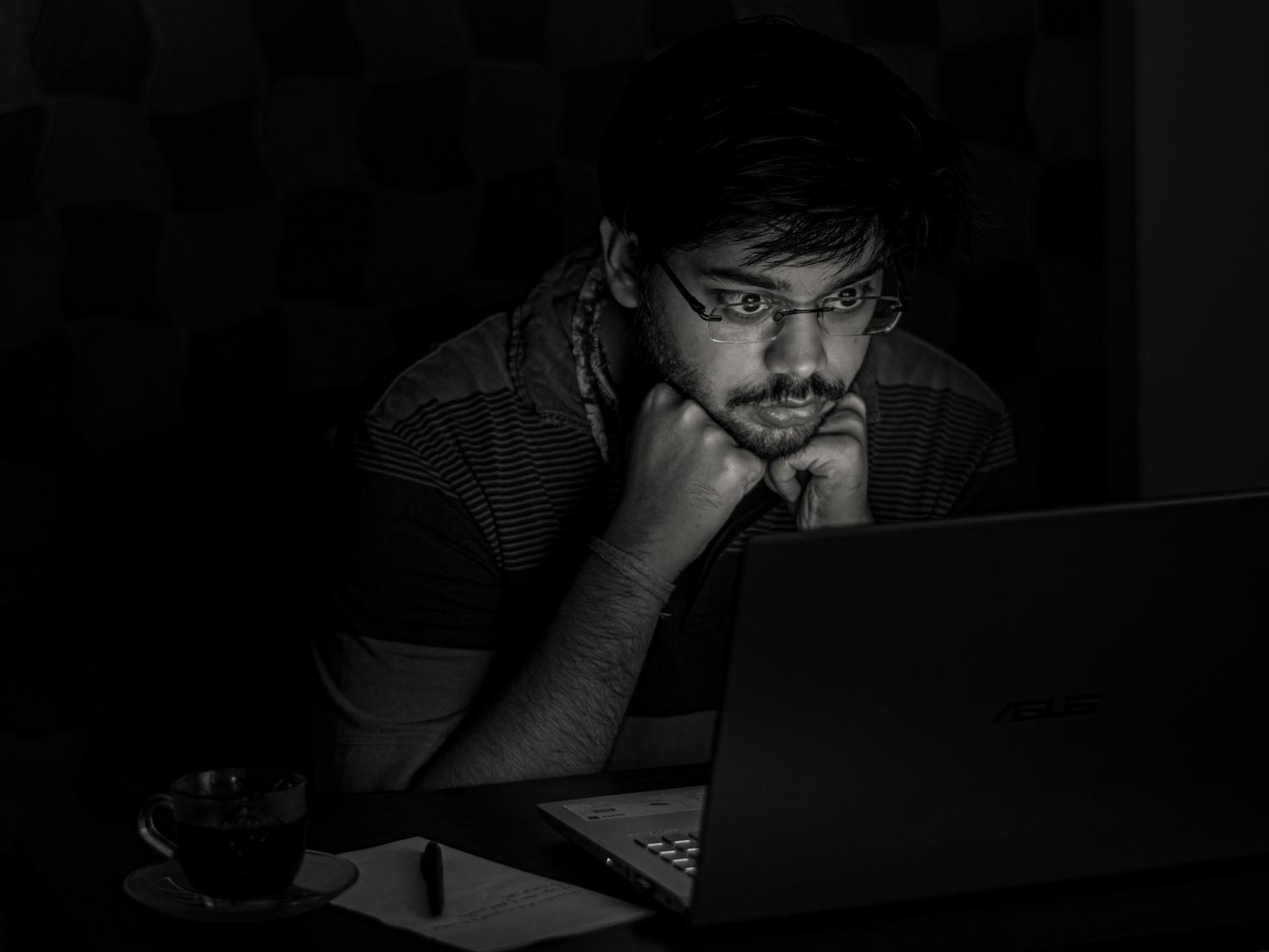 Hombre trabajando en una habitación oscura donde la pantalla del portátil es la única fuente de luz, ilustrando la fatiga por iluminación deficiente. Hombre trabajando en una habitación oscura donde la pantalla del portátil es la única fuente de luz, ilustrando la fatiga por iluminación deficiente.