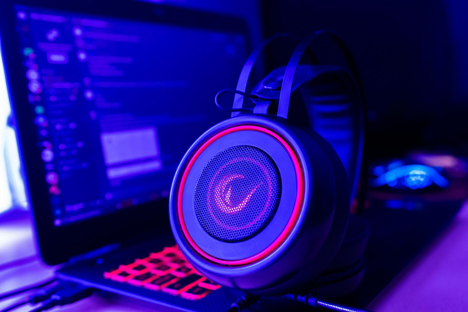 Detalle de unos auriculares gaming con iluminación LED roja y rosa junto a un ordenador portátil en un ambiente oscuro con luces azules. Detalle de unos auriculares gaming con iluminación LED roja y rosa junto a un ordenador portátil en un ambiente oscuro con luces azules.