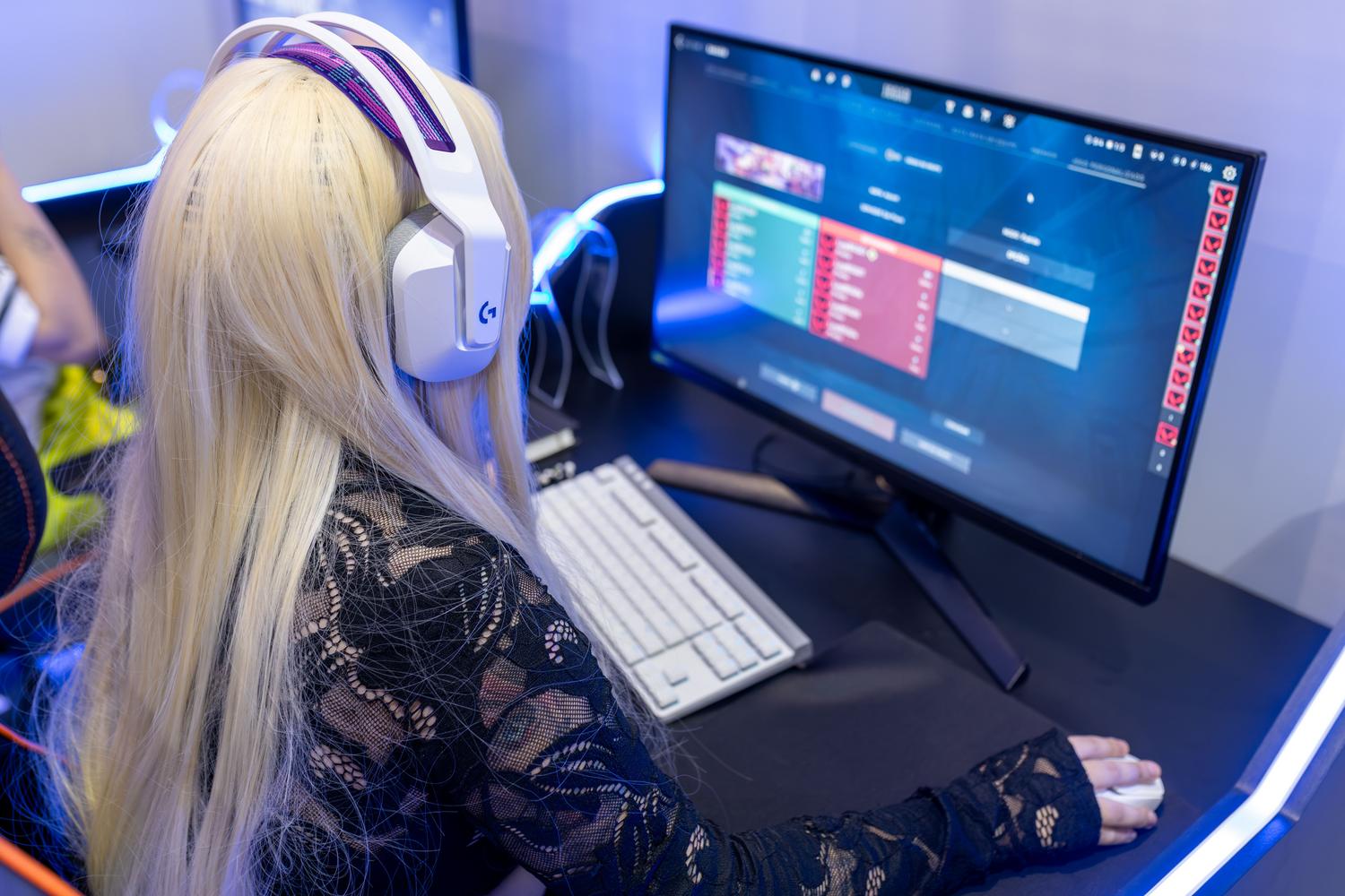 Vista trasera de una gamer con auriculares blancos y teclado mecánico en un setup moderno iluminado con tiras LED azules. Vista trasera de una gamer con auriculares blancos y teclado mecánico en un setup moderno iluminado con tiras LED azules.