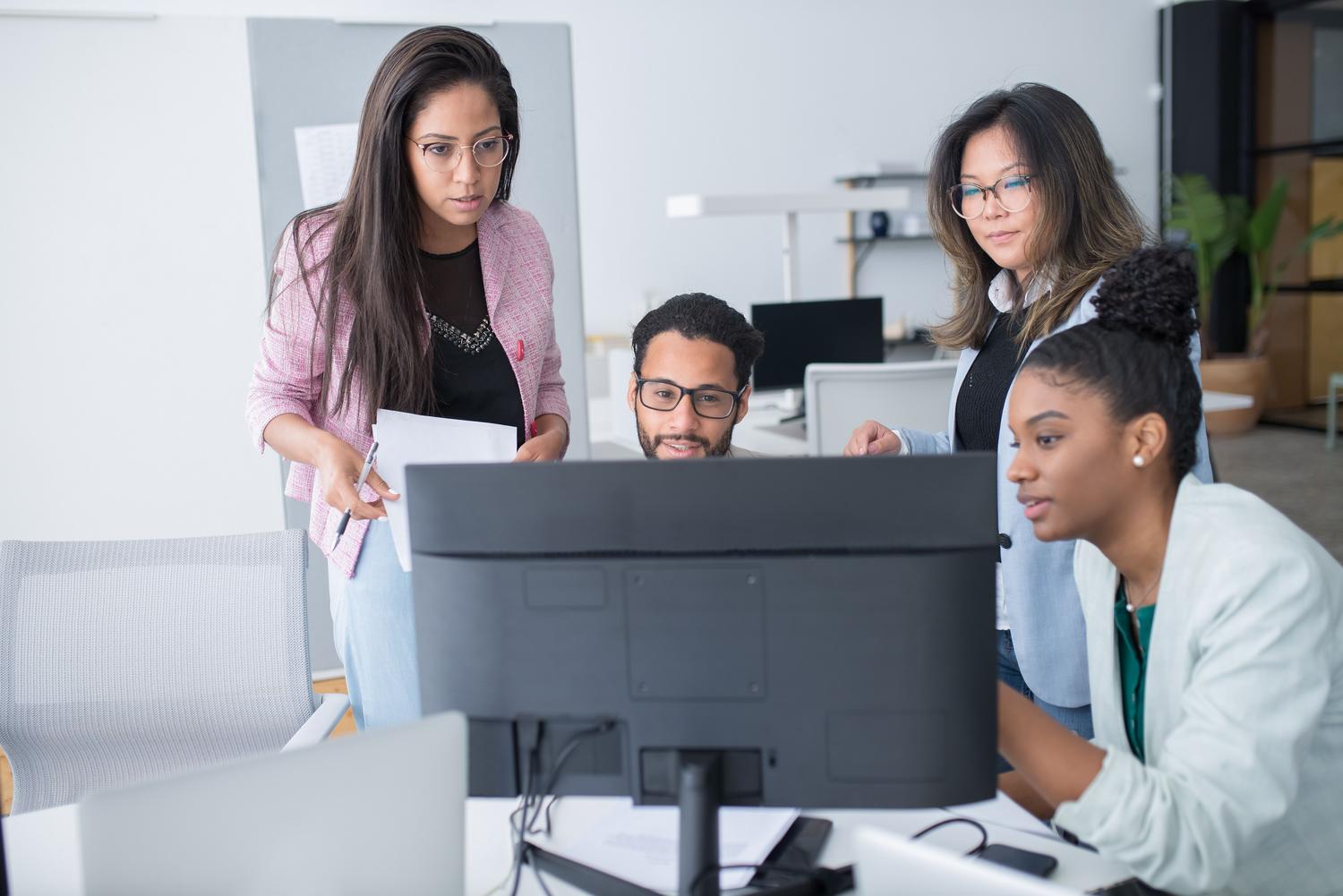Grupo diverso de profesionales colaborando estrechamente alrededor de un ordenador de escritorio en un espacio de trabajo profesional. Grupo diverso de profesionales colaborando estrechamente alrededor de un ordenador de escritorio en un espacio de trabajo profesional.