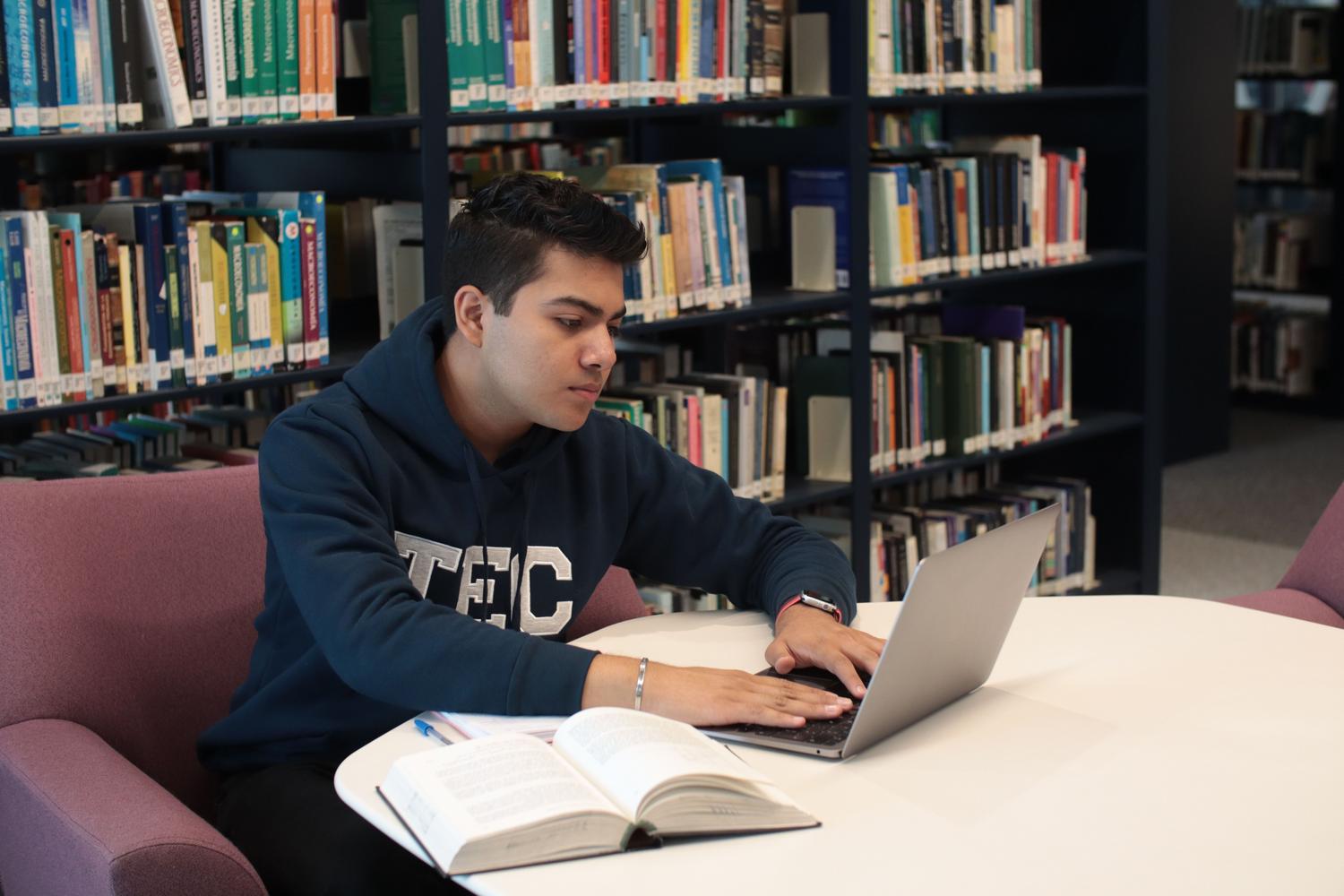 Estudiante universitario concentrado usando una laptop y revisando libros de texto en una biblioteca moderna. Estudiante universitario concentrado usando una laptop y revisando libros de texto en una biblioteca moderna.