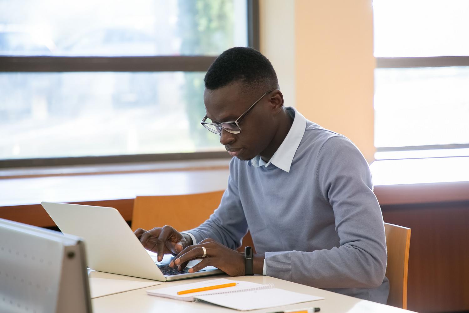 Joven estudiante con gafas concentrado escribiendo en su laptop mientras toma notas en una libreta. Joven estudiante con gafas concentrado escribiendo en su laptop mientras toma notas en una libreta.
