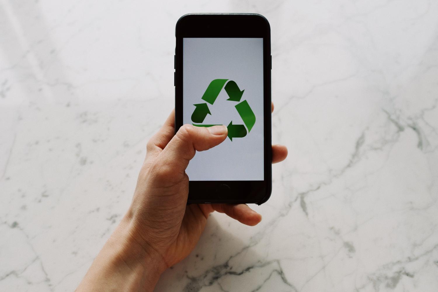 Mano sosteniendo un smartphone con un símbolo de reciclaje en pantalla, simbolizando la economía circular en la tecnología. Mano sosteniendo un smartphone con un símbolo de reciclaje en pantalla, simbolizando la economía circular en la tecnología.