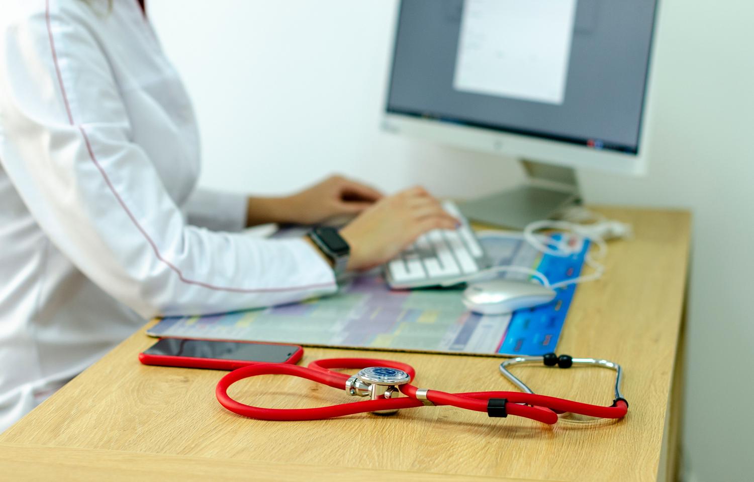 Médico trabajando en un ordenador con un estetoscopio al lado, representando la implementación de Historias Clínicas Electrónicas. Médico trabajando en un ordenador con un estetoscopio al lado, representando la implementación de Historias Clínicas Electrónicas.