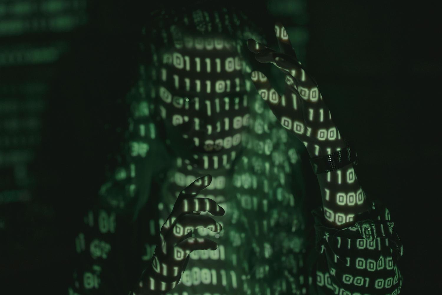 Persona con capucha y código binario proyectado sobre su cuerpo, representando la ciberseguridad, la auditoría de código y la identidad digital. Persona con capucha y código binario proyectado sobre su cuerpo, representando la ciberseguridad, la auditoría de código y la identidad digital.