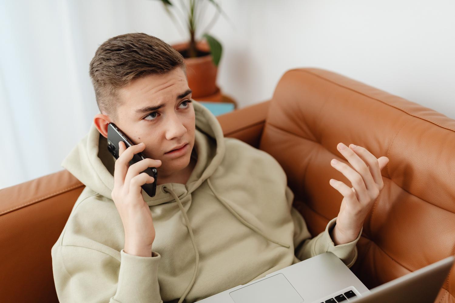 Hombre joven verificando información en su portátil mientras habla por teléfono, representando la verificación independiente. Hombre joven verificando información en su portátil mientras habla por teléfono, representando la verificación independiente.
